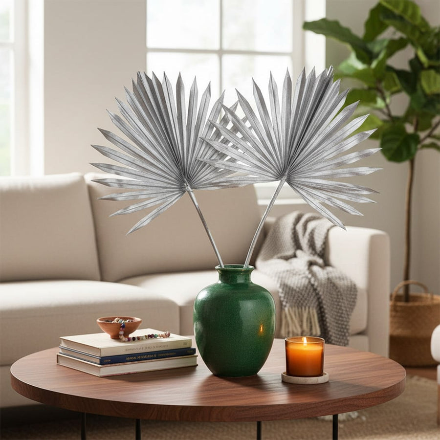 Green ceramic vase holding silver fan palm leaves placed on a round wooden coffee table with books candle and sofa in a warm modern living room.
