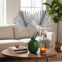 Green ceramic vase holding silver fan palm leaves placed on a round wooden coffee table with books candle and sofa in a warm modern living room.