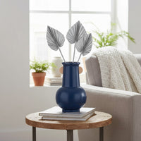 Deep blue ceramic vase with silver palm leaves styled on a small wooden side table beside a sofa in a bright modern living room.