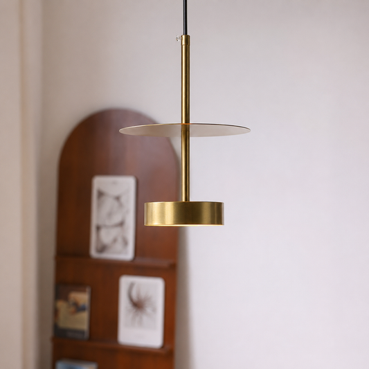 A modern brass pendant light with a minimalist two-tiered design, hanging against a neutral wall with a blurred wooden shelf in the background.