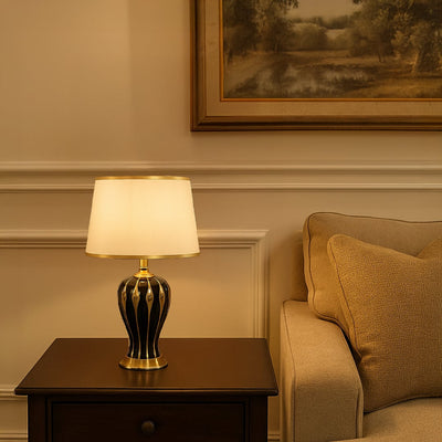 White shade dark ceramic table lamp on wooden side table with light on beside beige sofa for warm classic décor.