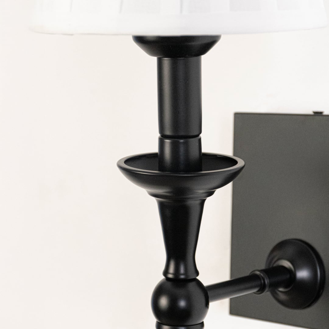 Closeup of black metal wall sconce with white pleated lampshade highlighting classic detailing and smooth finish.