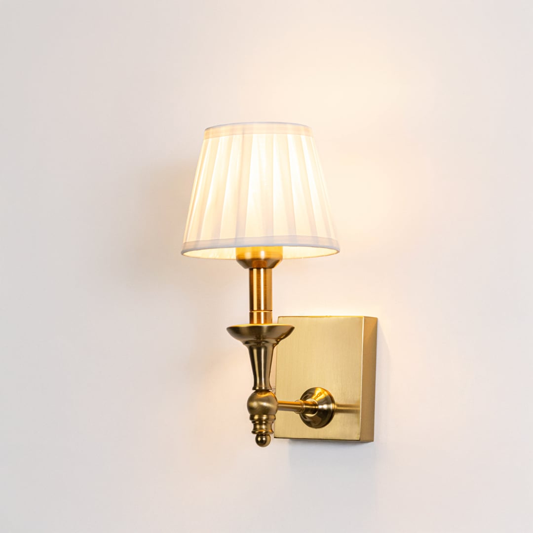 Gold wall sconce with white pleated shade illuminated on a bright white background for clear product visibility.