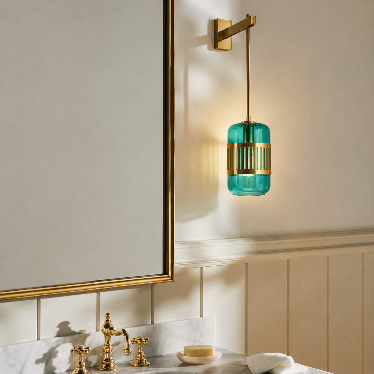A wall-mounted bathroom light fixture with a cylindrical teal glass shade and gold metal accents, shown installed above a marble vanity with gold fixtures in a real interior setting.