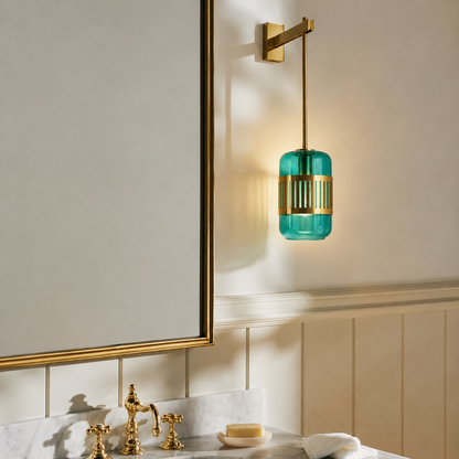 A wall-mounted bathroom light fixture with a cylindrical teal glass shade and gold metal accents, shown installed above a marble vanity with gold fixtures in a real interior setting.