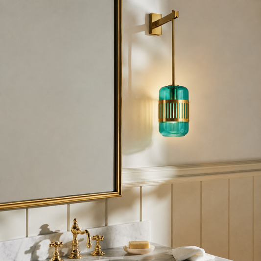 A wall-mounted bathroom light fixture with a cylindrical teal glass shade and gold metal accents, shown installed above a marble vanity with gold fixtures in a real interior setting.