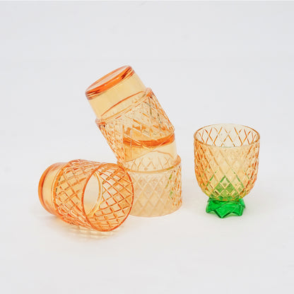 Four translucent orange glasses with diamond-cut patterns, including two stacked on top of each other, one lying on its side, and one standing upright with a green leaf-shaped base, photographed on a clean white background.