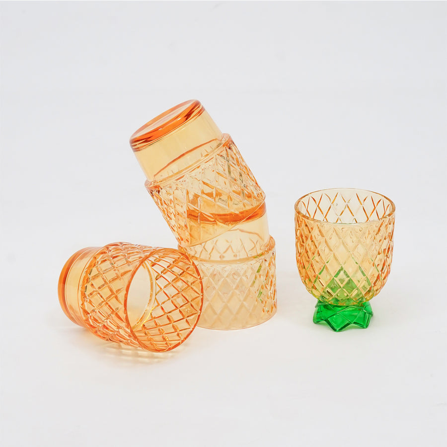 Four translucent orange glasses with diamond-cut patterns, including two stacked on top of each other, one lying on its side, and one standing upright with a green leaf-shaped base, photographed on a clean white background.