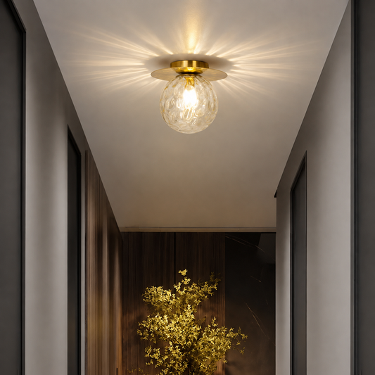 A modern ceiling light fixture with a gold brass circular canopy and a textured clear glass globe bulb, emitting a warm starburst glow in a hallway setting.
