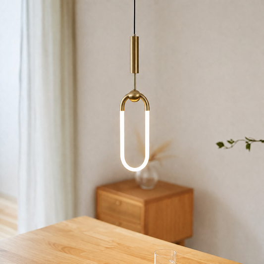 A modern pendant light with a glowing white U-shaped tube and a brushed brass-colored top and middle section, hanging in a softly lit room above a wooden table.