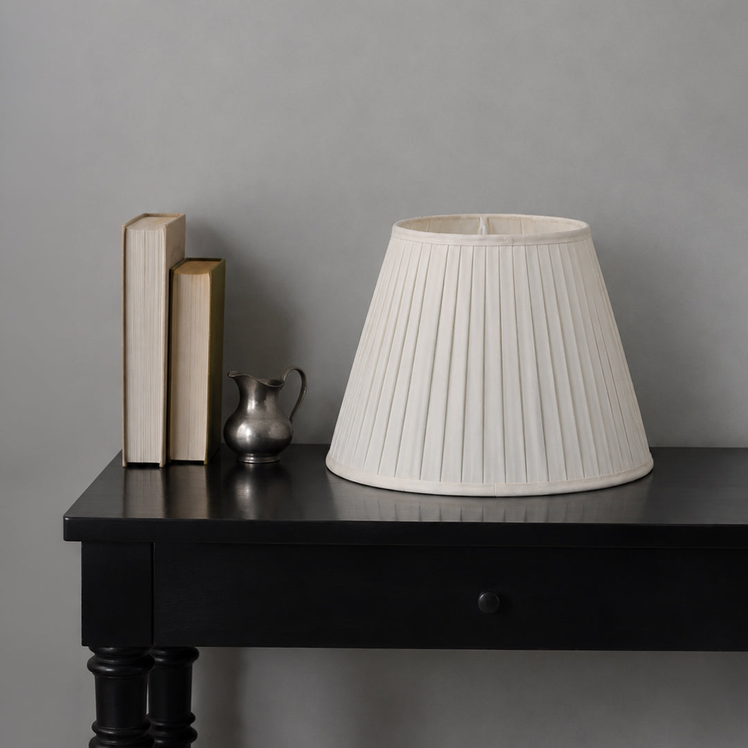 An off-white pleated fabric lampshade displayed on a black wooden table next to decorative books and a small silver pitcher against a neutral gray wall.