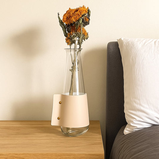 Minimal glass decorative vase with beige metal sleeve placed on wooden bedside table holding dried orange flowers for modern bedroom home decor.