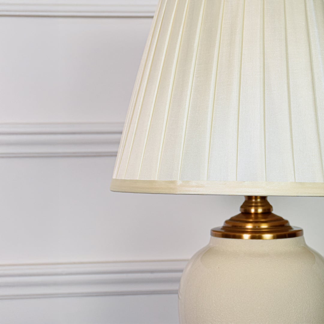 Closeup of ivory ceramic table lamp showing smooth glazed surface curved form gold metal base and fabric shade texture.