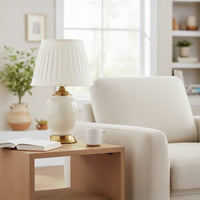 Ivory ceramic table lamp with smooth glazed body gold metal base and pleated fabric shade styled on bedside or side table.