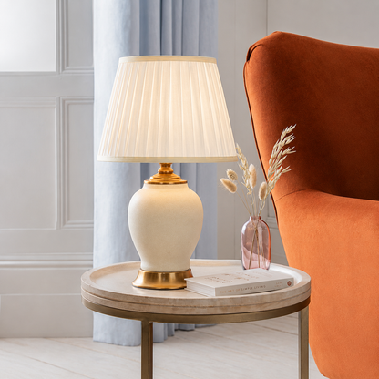 Elegant table lamp with a white pleated shade on a round side table beside an orange accent chair in a modern living room.