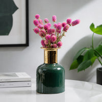 Dark green glossy vase with gold neck and pink flowers on white table for vibrant luxury home interior decor.