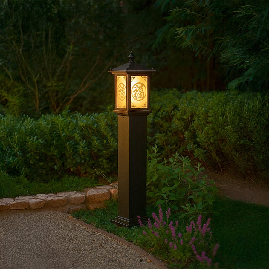 Brown outdoor pillar lamp with glowing decorative Chinese pattern glass lighting a garden pathway at night creating a warm inviting ambience among lush greenery.