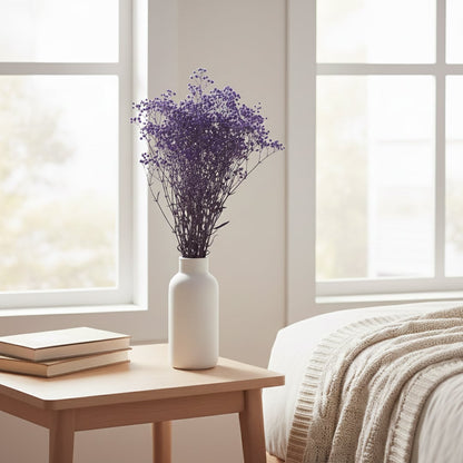 A vibrant purple dried glixia flower bouquet in a white vase on a wooden bedside table in a cozy sunny bedroom setup.