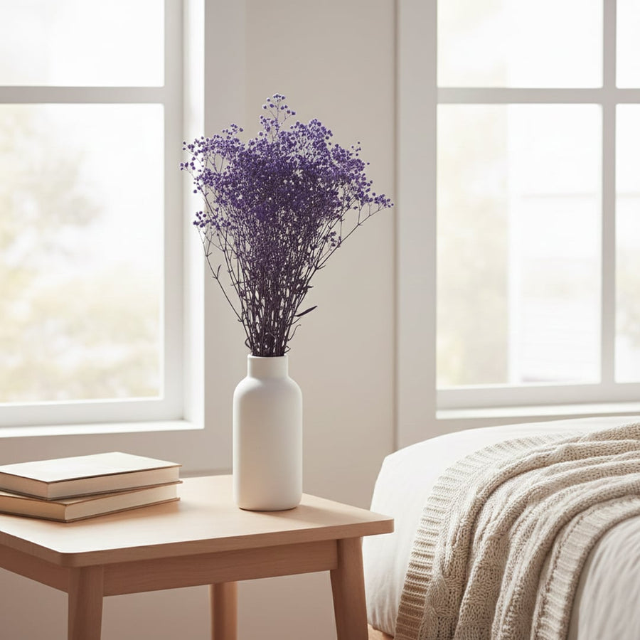 A vibrant purple dried glixia flower bouquet in a white vase on a wooden bedside table in a cozy sunny bedroom setup.