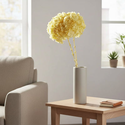 Yellow preserved hydrangea flowers in a tall white vase on a wooden side table next to a living room couch in daylight.