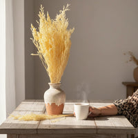 Bright yellow dried ornamental grass stalks in a ceramic vase on a rustic wooden dining table next to a steaming coffee cup.