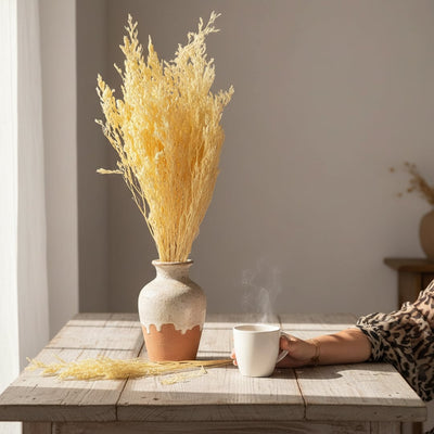 Bright yellow dried ornamental grass stalks in a ceramic vase on a rustic wooden dining table next to a steaming coffee cup.