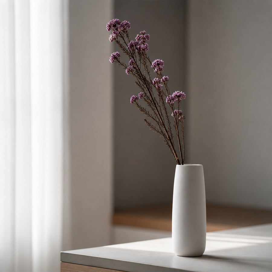 Dark purple dried wildflower stems arranged inside a sleek modern white ceramic vase on a windowsill in soft daylight.