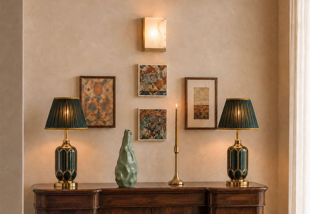 A styled interior scene with two matching table lamps on a dark wood console against a beige wall, flanking a vase of branches and a candle. Above the console are four framed artworks and a wall sconce.
