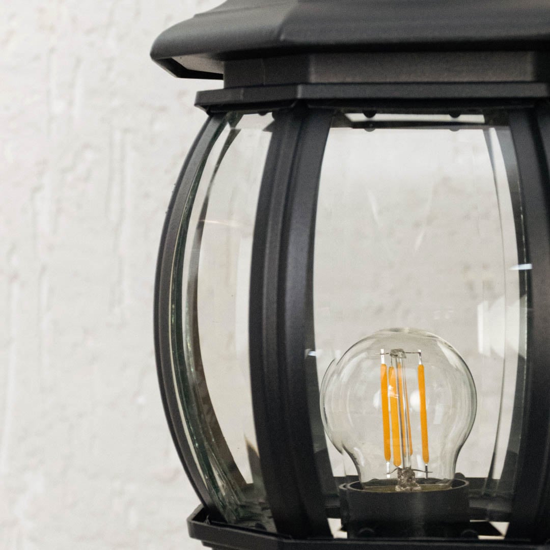 Detailed zoom view of black outdoor pillar lamp with curved transparent glass panels highlighting glowing LED bulb.