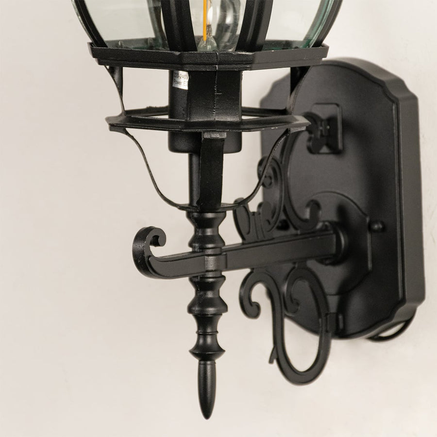 Closeup of black upward wall lantern showing curved glass cage detail decorative scroll bracket and ornate finial design.