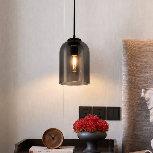 A modern industrial-style pendant light with a dark gray smoked glass dome shade and a visible Edison bulb, hanging in a bedroom setting above a bedside table.