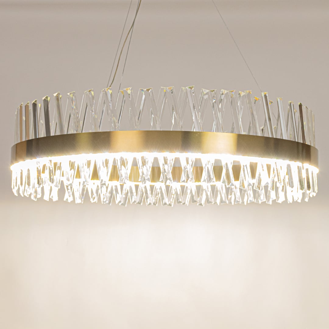 Gold metal chandelier with crystal rods shown with lights on creating a bright elegant and luxurious glow for modern interiors.