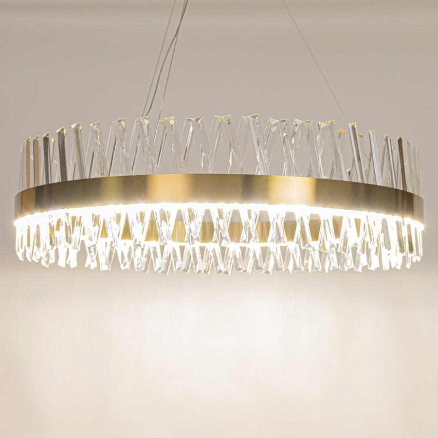 Gold metal chandelier with crystal rods shown with lights on creating a bright elegant and luxurious glow for modern interiors.