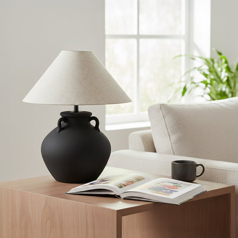 Matte black ceramic table lamp with sculptural pot style base and natural fabric shade styled on living room side table.