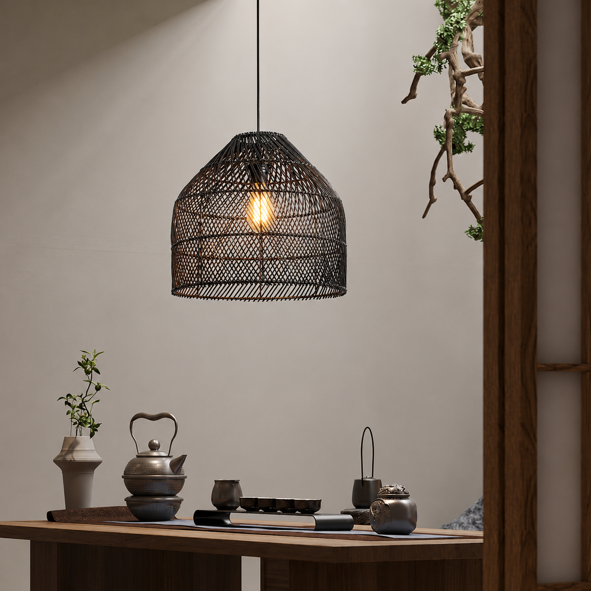 A black woven rattan pendant light hangs over a wooden table in a styled interior setting with tea accessories and plants.