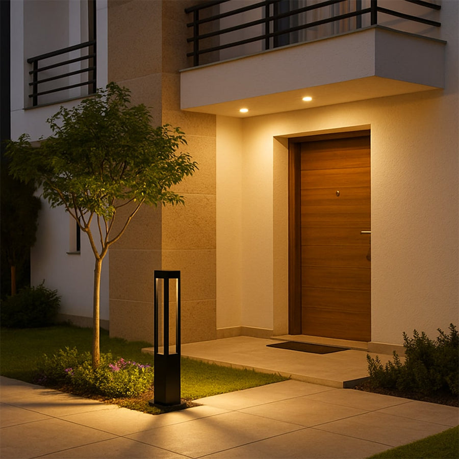 A modern outdoor bollard light glowing warmly beside a contemporary home entrance with soft illumination enhancing the pathway and garden area at night.