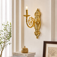 An ornate gold wall sconce with a single candle-style bulb, mounted on a cream-colored wall in a styled interior setting with a plant, books, and a framed picture.