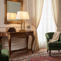 Modern gold table lamp with white fabric shade placed on an ornate wooden console beside a framed photo and books in a classic living room setting.