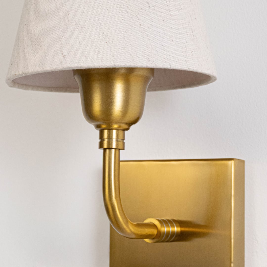 Closeup of gold metal wall light arm with fabric shade mount showing smooth finish refined curves and elegant lighting detail.