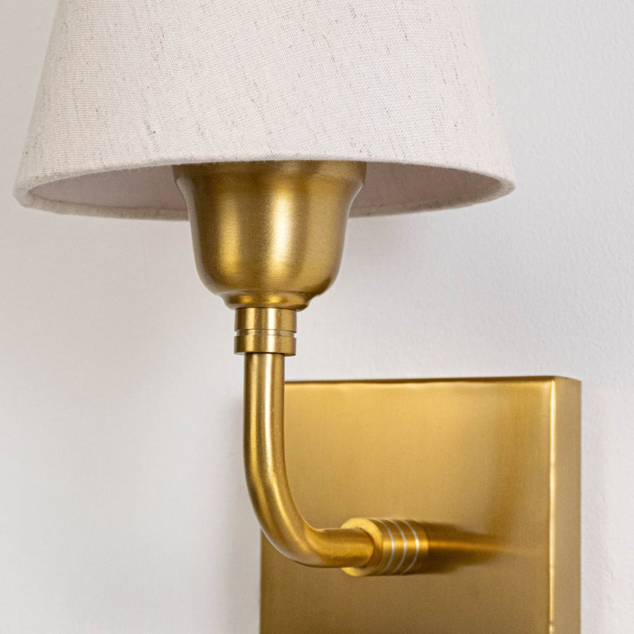 Closeup of gold metal wall light arm with fabric shade mount showing smooth finish refined curves and elegant lighting detail.