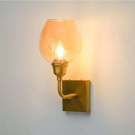 A wall-mounted sconce with a warm brass-finished metal bracket and a clear glass shade glowing with a yellow light, mounted on a plain light gray wall.