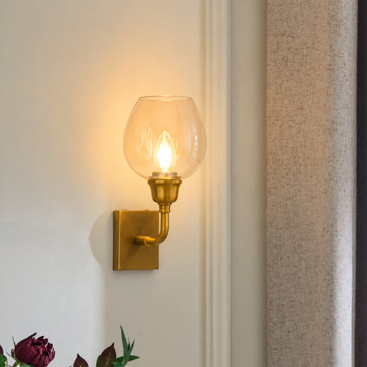 A wall-mounted sconce with a clear glass globe shade and a gold-finished metal fixture, lit against an off-white wall in an interior setting.