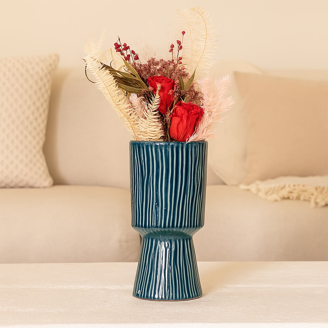 A tall blue textured vase displayed on a soft beige sofa setup featuring red roses and pastel dried stems creating a warm and elegant home styling look.