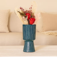 A tall blue textured vase displayed on a soft beige sofa setup featuring red roses and pastel dried stems creating a warm and elegant home styling look.