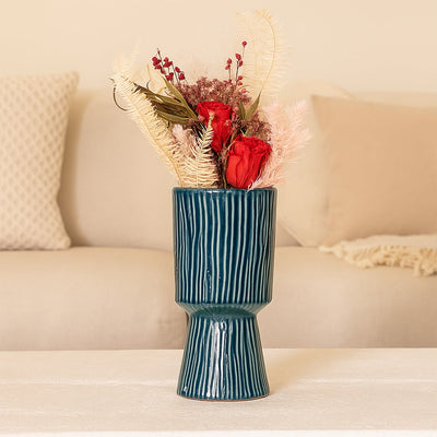 A tall blue textured vase displayed on a soft beige sofa setup featuring red roses and pastel dried stems creating a warm and elegant home styling look.