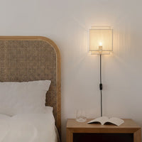 Minimalist white pleated fabric wall sconce casting warm light over a cozy bedroom wooden nightstand and cane bedstead.