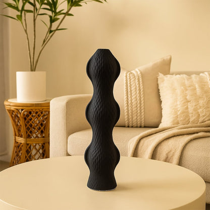 Tall black sculptural vase with a wave-like pattern displayed on a beige round table in a cozy living room with soft-toned decor and textured cushions.