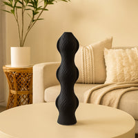 Tall black sculptural vase with a wave-like pattern displayed on a beige round table in a cozy living room with soft-toned decor and textured cushions.