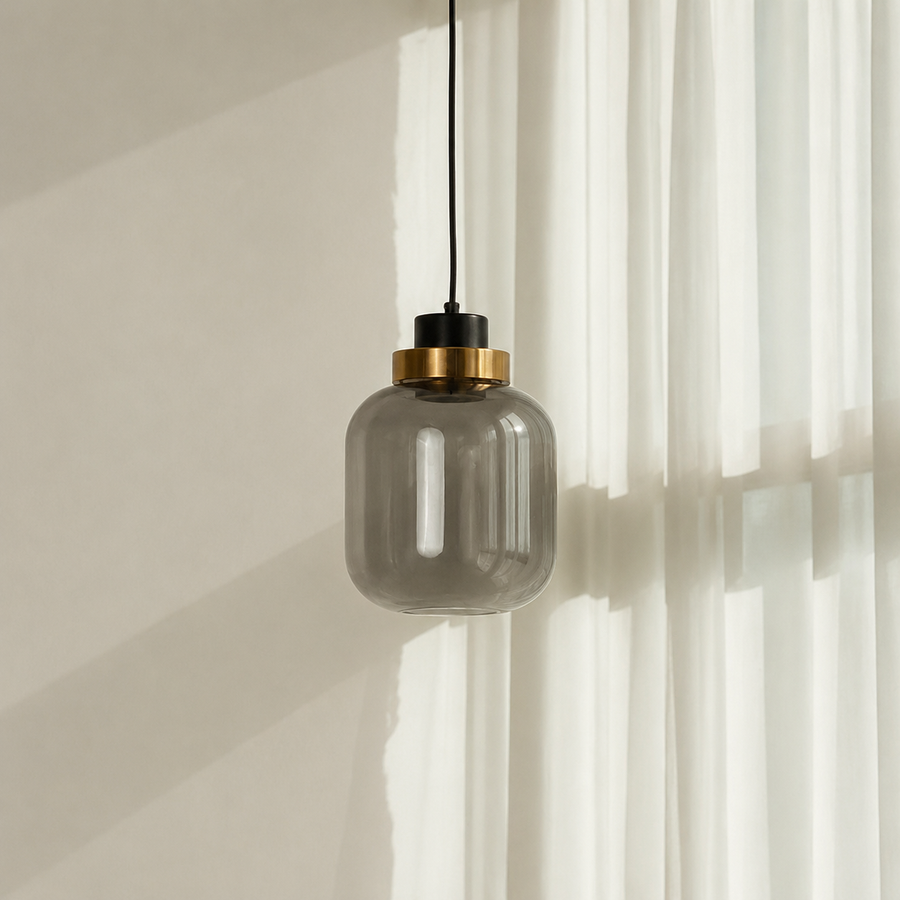 A modern pendant light with a smoky translucent glass shade, a black top cap, and a brushed brass ring, hanging against a warm beige wall with white sheer curtains.