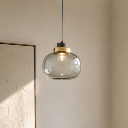 A modern pendant light with a clear glass dome shade, a brushed brass metal collar, and a black ceiling mount, hanging in a minimalist room with a textured beige wall.